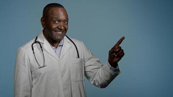 African American man doctor medical worker in white coat male healthcare employee medic customer in studio blue background pointing index finger making choice choosing smiling good selection medicare photo