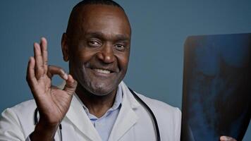 Close-up mature african american man doctor surgeon traumatologist radiologist in medical coat in studio blue background hold x-ray review result tomogram showing sign ok gesture approval good health photo