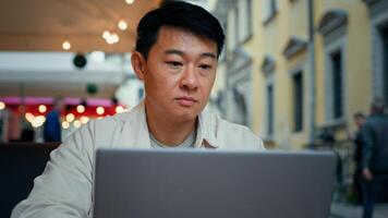 Close-up asian man sitting in cafe typing on laptop chatting online reading good message male tourist browses social network chooses hotel on internet communicates on dating site enjoys virtual chat photo