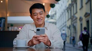 Smiling asian man tourist sitting in street cafe holding mobile phone booking tickets online remotely communicates with friends in social network on smartphone using free wifi makes order on internet photo