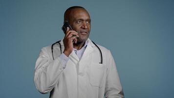 Mature african american man doctor talking on phone with patient in blue background studio male therapist in medical gown consult client using smartphone remote medical assistance healthcare concept photo