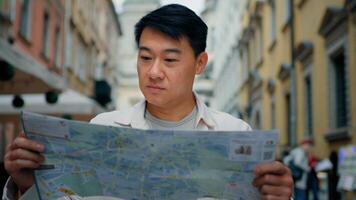 Asian male tourist traveling in new city standing outdoors looking at paper map in search of location using travel guide to get directions young man traveler international student searching right way photo