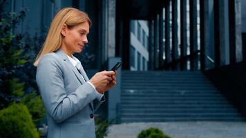 Focused successful businesswoman walking outdoors holding smartphone writing email message communicating with friend or client on social media using mobile phone making order in online store side view photo