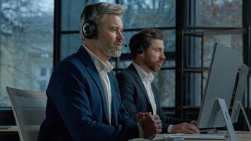 Two men call center service operators work in office talk to client using headset on computer concentrated sales agents sell online answering incoming call provide professional support to customers photo