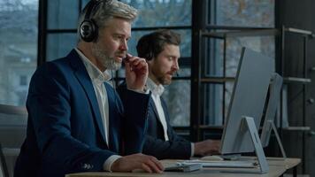 Two men call center service operators work in office talk to client using headset on computer concentrated sales agents sell online answering incoming call provide professional support to customers photo