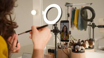 Glad Caucasian woman blogger girl influencer filming mobile phone on stabilizer lamp studio female vlogger streamer talk demonstrating shadow palette do makeup tutorial beauty stream blog mirror photo