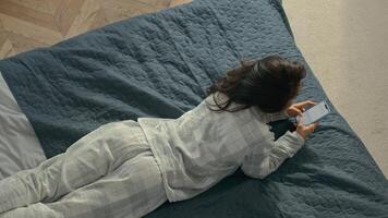Top view young cheerful brunette woman in pajamas lying on bed cozy comfortable home inside bedroom female roll over chatting texting smartphone mobile phone telephone communicate smiling weekend joy photo