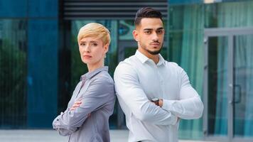 Two confident successful serious multiethnic business co-workers multiracial colleagues spaniard man and caucasian woman standing outdoors near city building crossing arms posing looking at camera photo
