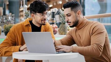Two arab men sit looking at laptop screen communicate smiles studying using new computer application browsing web pages online dating setting up software young guy explaining to friend helping support photo