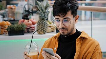 Young happy guy sits in cafe drinks freshly squeezed juice satisfied man holding mobile phone typing message email communicate with friends remote in social network online uses free wifi on smartphone photo