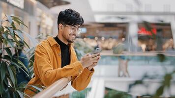 Young hispanic guy stands in mall holding phone smiling sending email on social network surfing web page checks news uses mobile application on smartphone making order in online store on telephone photo