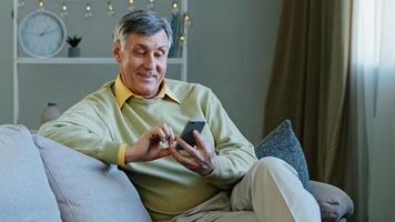 Caucasian elderly adult man sitting on sofa at home with gadget using online app on smartphone makes online order old grandfather resting reading news in mobile phone browsing scrolling websites photo