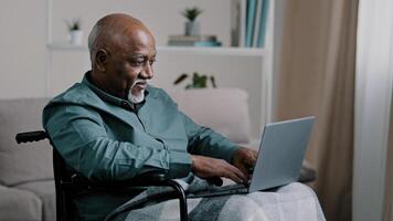 Old 60s african business man in wheelchair indoor at home typing on laptop communicates remotely working online distant texting chatting with family grandfather with disability use modern computer app photo