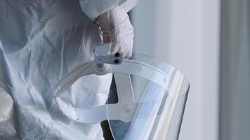 Unrecognizable doctor medical worker nurse practitioner scientist stands in clinic laboratory hospital at window wears white uniform and latex gloves holds in hand transparent protective face shield photo