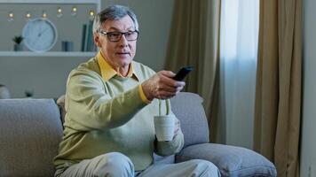 Grey-haired aged man in glasses sitting on couch watching TV enjoying relaxing at home drinking tea coffee aged male uses remote control to switch channels smiling enjoy funny movie television program photo