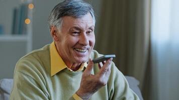 Mature grey-haired 60s man 70s senior old male at home hold smartphone speak loud to phone microphone speaker audio voice message using speakerphone talking with children remote mobile conversation photo