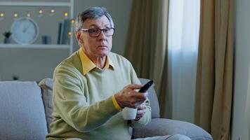 Caucasian aged senior mature man in glasses relax on couch at home change channel with remote controller watching TV program relaxing with drinking tea coffee old male choose movie television indoors photo