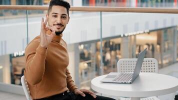 Young arab guy student freelancer typing on laptop surfing social network on internet admiring work new application showing approval sign consent gesture ok smiles looking at camera approving software photo