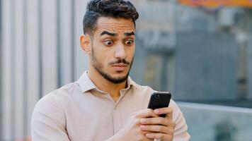 Portrait bearded young amazed handsome business man stands in city looking in mobile phone with shocked expression on face reads bad news good surprised browsing web receiving message shock fright photo