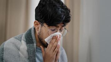 Close-up sad exhausted arabic bearded man suffering from seasonal allergy dust virus respiratory disease runny nose guy in glasses wrapped in blanket sitting at home blowing nose snot in paper napkin photo