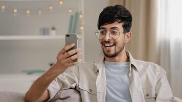 Portrait indoor male arabic guy unshaven man with glasses sitting on couch at home chatting online using mobile phone reads news scrolling browsing net typing in smartphone surprised answers message photo
