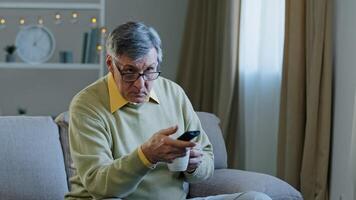 Caucasian aged senior man in glasses sitting on couch change channel with remote controller watching TV relaxing at home drinking tea coffee old male choose movie television program at home resting photo