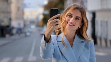 Portrait of young beautiful girl Caucasian business woman blogger vlogger model stands in city holds phone taking photo on mobile camera shoots for vlog blog with smartphone online chat call