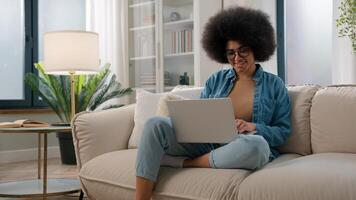 Smiling African American woman on couch at home using laptop smiling online work studying distance typing message happy lady girl chatting on computer browsing internet social media watch funny photo
