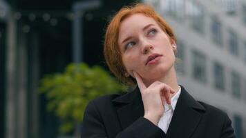 Close up young puzzled Caucasian ginger businesswoman girl female woman student elegant suit think finger touch cheek contemplating gesture outdoors city street. Business choice decision idea planning photo