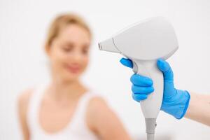 A young, happy woman is relaxed in a modern laser hair removal clinic. She sits against a light background, with a laser device handle visible nearby photo