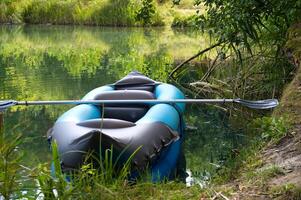 Inflatable kayak with paddle resting on serene lakeshore surrounded by lush green trees photo