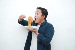 A portrait of happy Asian man wearing a blue shirt while holding a plate of fried noodles. Young man smiling and pointing to a plate of noodle. Isolated with a white background. photo