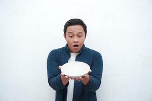 Portrait of happy Asian man wearing a blue shirt while holding an empty plate with happy and excited expression. Young man showing and pointing empty mockup plate. Isolated with a white background. photo