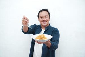 A portrait of happy Asian man wearing a blue shirt while holding a plate of fried noodles. Young man smiling and pointing to a plate of noodle. Isolated with a white background. photo