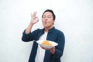 A portrait of happy Asian man wearing a blue shirt while holding a plate of fried noodles. Young man smiling and pointing to a plate of noodle. Isolated with a white background. photo