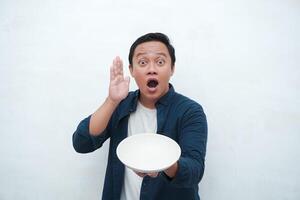 Portrait of happy Asian man wearing a blue shirt while holding an empty plate with happy and excited expression. Young man showing and pointing empty mockup plate. Isolated with a white background. photo