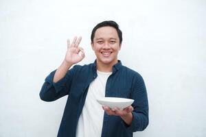 Portrait of happy Asian man wearing a blue shirt while holding an empty plate with happy and excited expression. Young man showing and pointing empty mockup plate. Isolated with a white background. photo