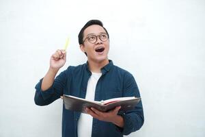 Happy young Asian man with glasses showing expression of gets genius idea. Asian college student showing happy and excited expression while holding book and pen photo