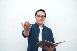 Young Asian man holding books smiling happy and positive while reading a book. Asian student pointing and showing welcoming gesture photo