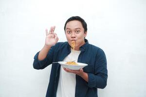 A portrait of happy Asian man wearing a blue shirt while holding a plate of fried noodles. Young man smiling and pointing to a plate of noodle. Isolated with a white background. photo