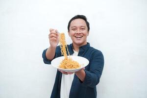 A portrait of happy Asian man wearing a blue shirt while holding a plate of fried noodles. Young man smiling and pointing to a plate of noodle. Isolated with a white background. photo