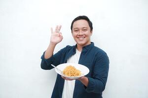 A portrait of happy Asian man wearing a blue shirt while holding a plate of fried noodles. Young man smiling and pointing to a plate of noodle. Isolated with a white background. photo