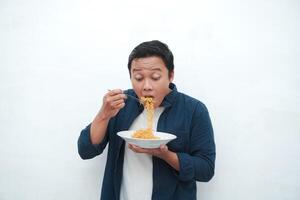 A portrait of happy Asian man wearing a blue shirt while holding a plate of fried noodles. Young man smiling and pointing to a plate of noodle. Isolated with a white background. photo
