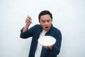 Portrait of happy Asian man wearing a blue shirt while holding an empty plate with happy and excited expression. Young man showing and pointing empty mockup plate. Isolated with a white background. photo