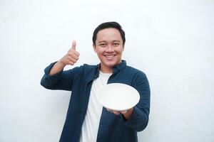 Portrait of happy Asian man wearing a blue shirt while holding an empty plate with happy and excited expression. Young man showing and pointing empty mockup plate. Isolated with a white background. photo