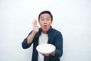 Portrait of happy Asian man wearing a blue shirt while holding an empty plate with happy and excited expression. Young man showing and pointing empty mockup plate. Isolated with a white background. photo