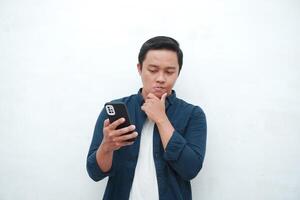 Young Asian man thinking and concentrating while holding books and handphone. Asian student thinking and looking for ideas while holding phone over white background photo