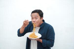 A portrait of happy Asian man wearing a blue shirt while holding a plate of fried noodles. Young man smiling and pointing to a plate of noodle. Isolated with a white background. photo
