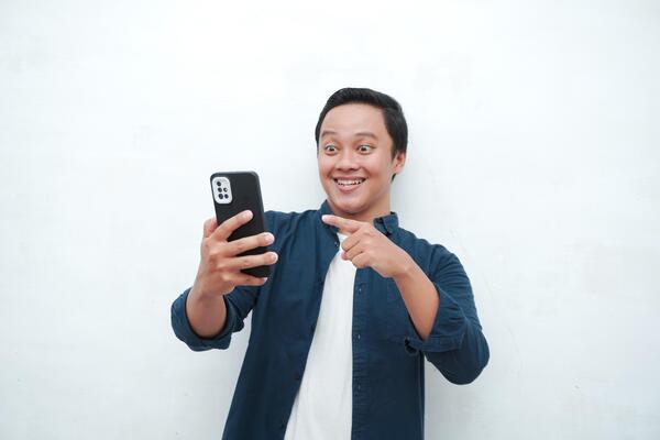 Young Asian man looking smartphone with shocked and surprised expression. Male with blue t-shirt showing excited expression isolated on white background