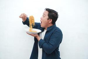A portrait of happy Asian man wearing a blue shirt while holding a plate of fried noodles. Young man smiling and pointing to a plate of noodle. Isolated with a white background. photo